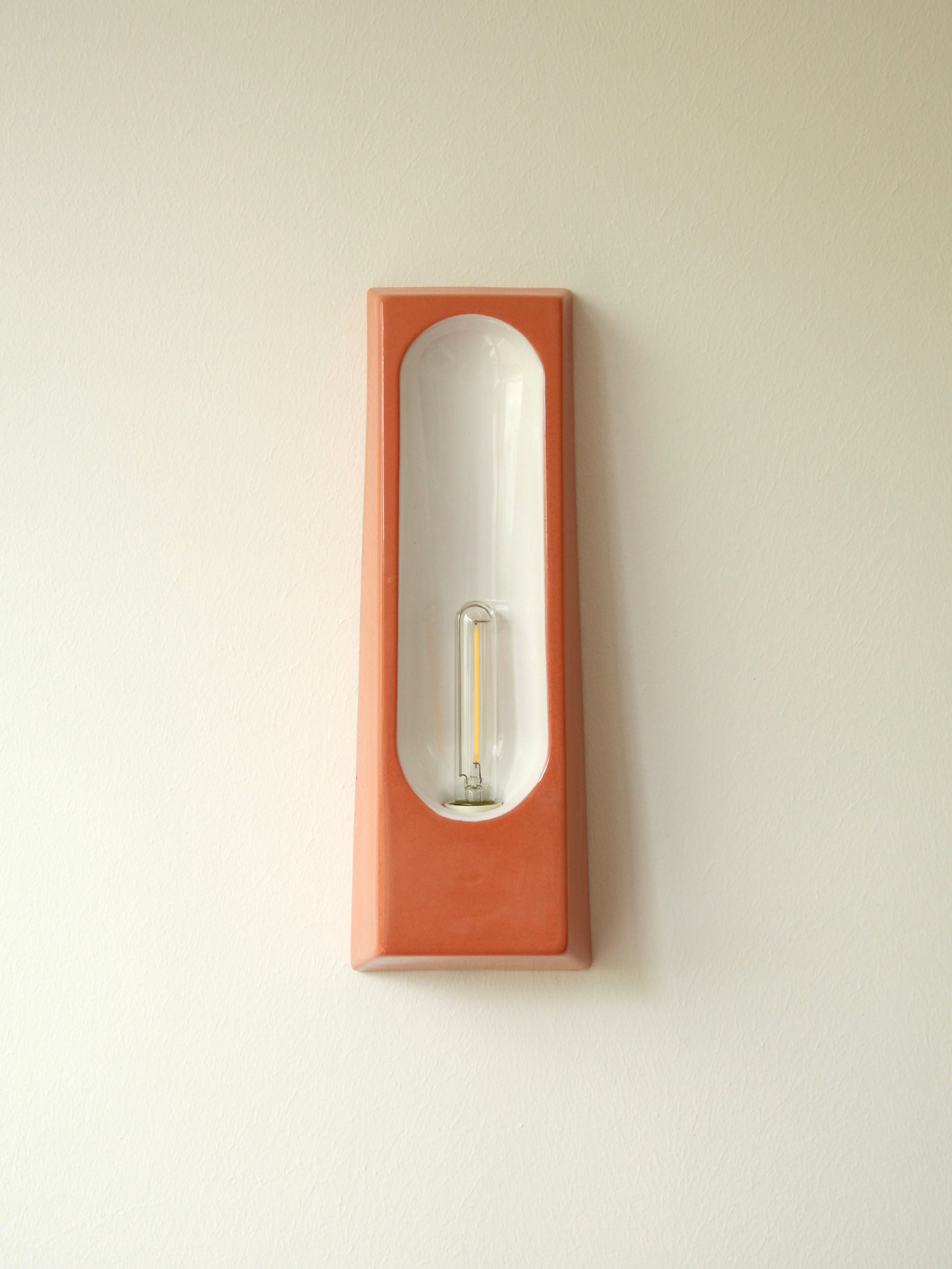 The Violaine d'Harcourt Alcove Terra B Glossy is a glazed ceramic, terracotta-colored, rectangular wall-mounted light fixture with a clear elongated bulb that contrasts beautifully against its white backdrop on an off-white wall.