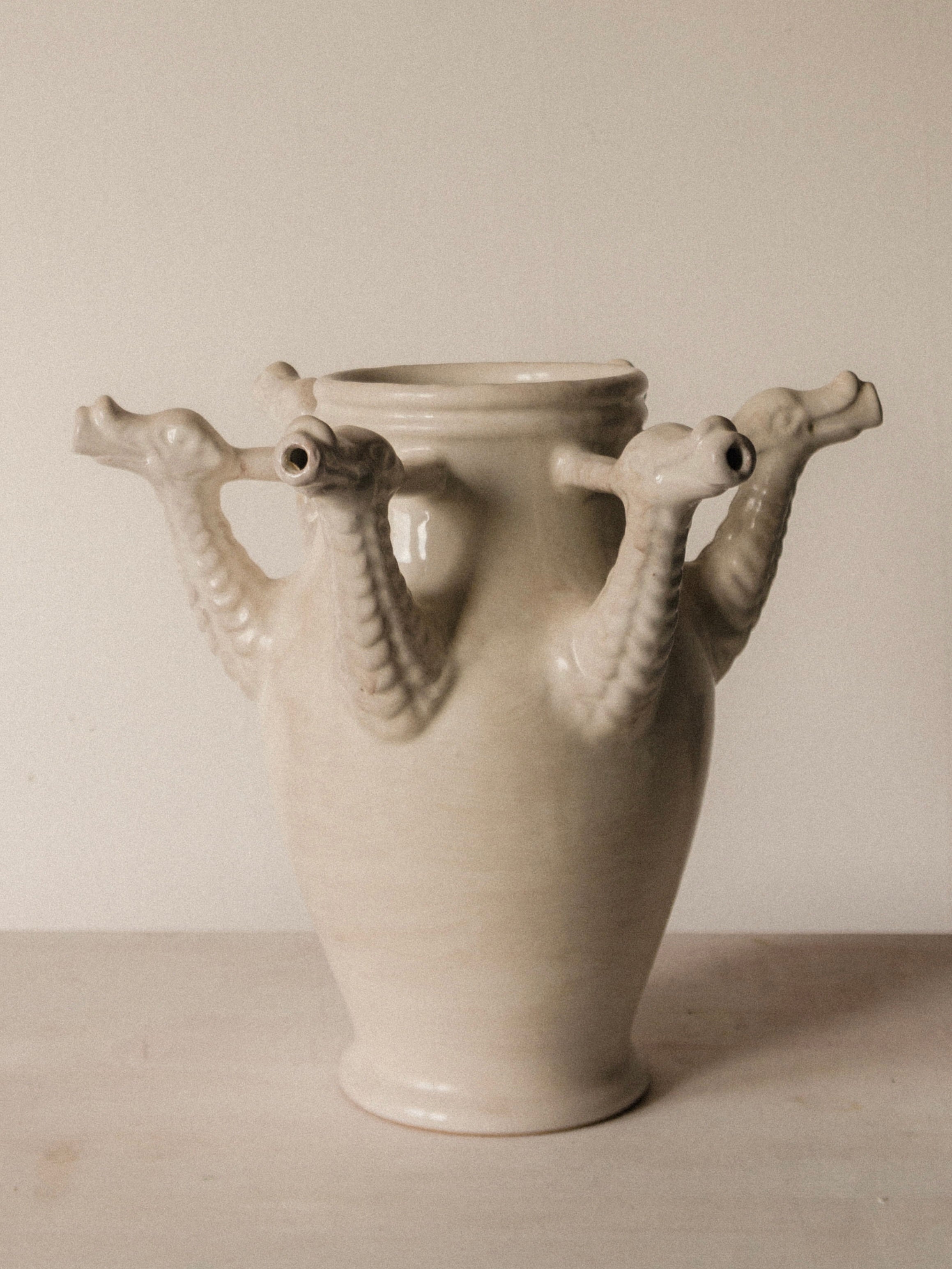 The Drago Bianco Vase by COSEINCORSO is a cream ceramic piece handmade in Italy, inspired by Castelli, with four sculpted dragon heads with open mouths extending from its sides, displayed against a plain light background.