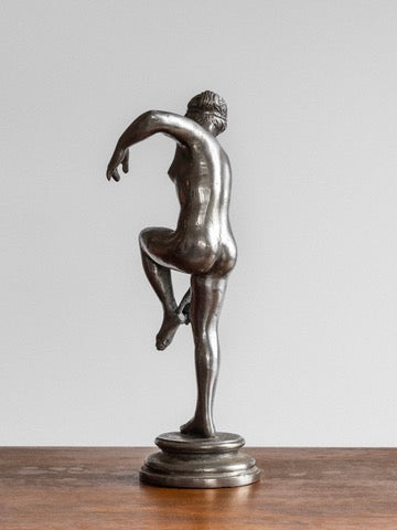 The Spigel Elegant Metal Sculpture, 1930s, features a bronze statue of a nude figure balancing on one leg with the other bent at the knee. The dynamic pose showcases one arm extended downward and the other outward. Reminiscent of 1930s Swedish sculpture, this piece is set on a round base placed on a wooden surface against a plain background.