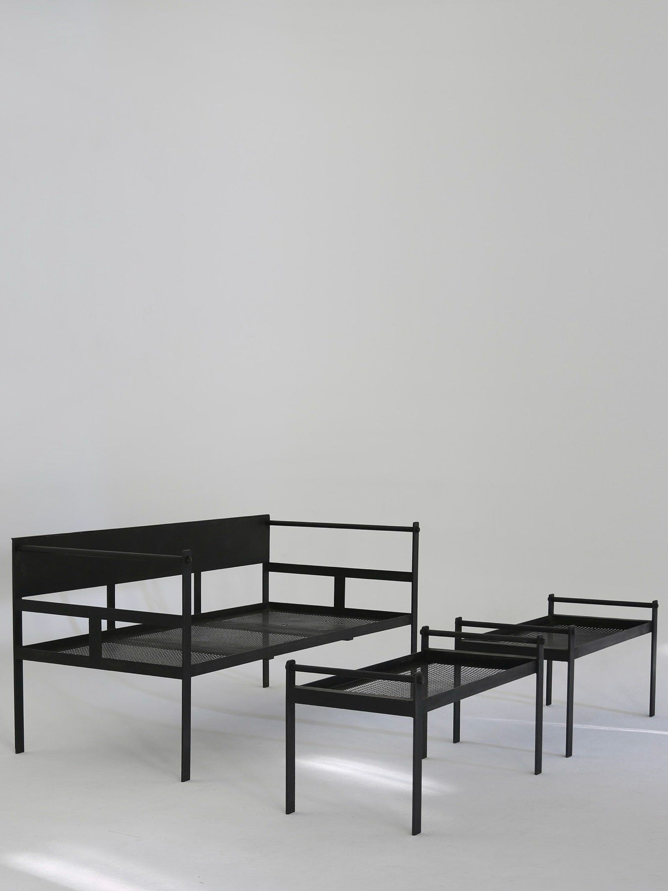 The Modernist Lounge Set by Ancien et Jolie features a minimalist black metal daybed with a geometric design on a light gray background, resembling Mathieu Mategot's style. The steel frame and perforated platform highlight its sleek lines and simplicity without a mattress.
