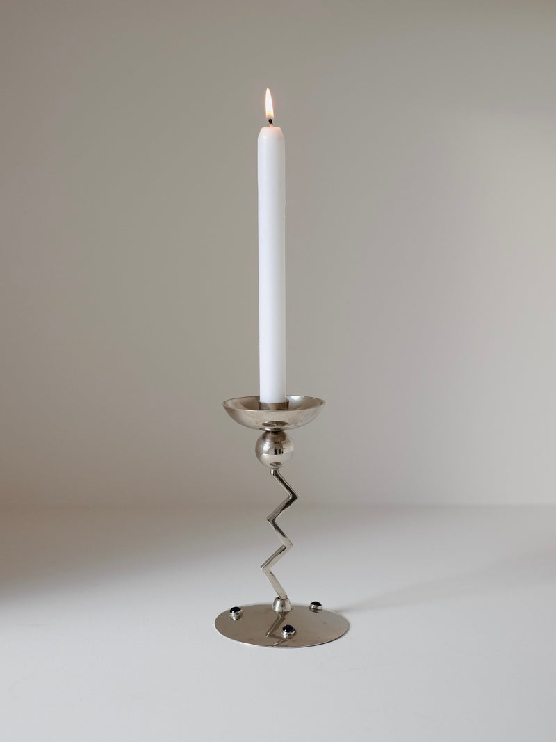 A lit candle in the Zigzag Candleholder by septembre studios sits on a flat surface. The plain, softly lit background enhances its Memphis-style design with cobalt blue inset stones and a metallic finish, highlighting the simple yet elegant charm of the 80's-inspired piece.