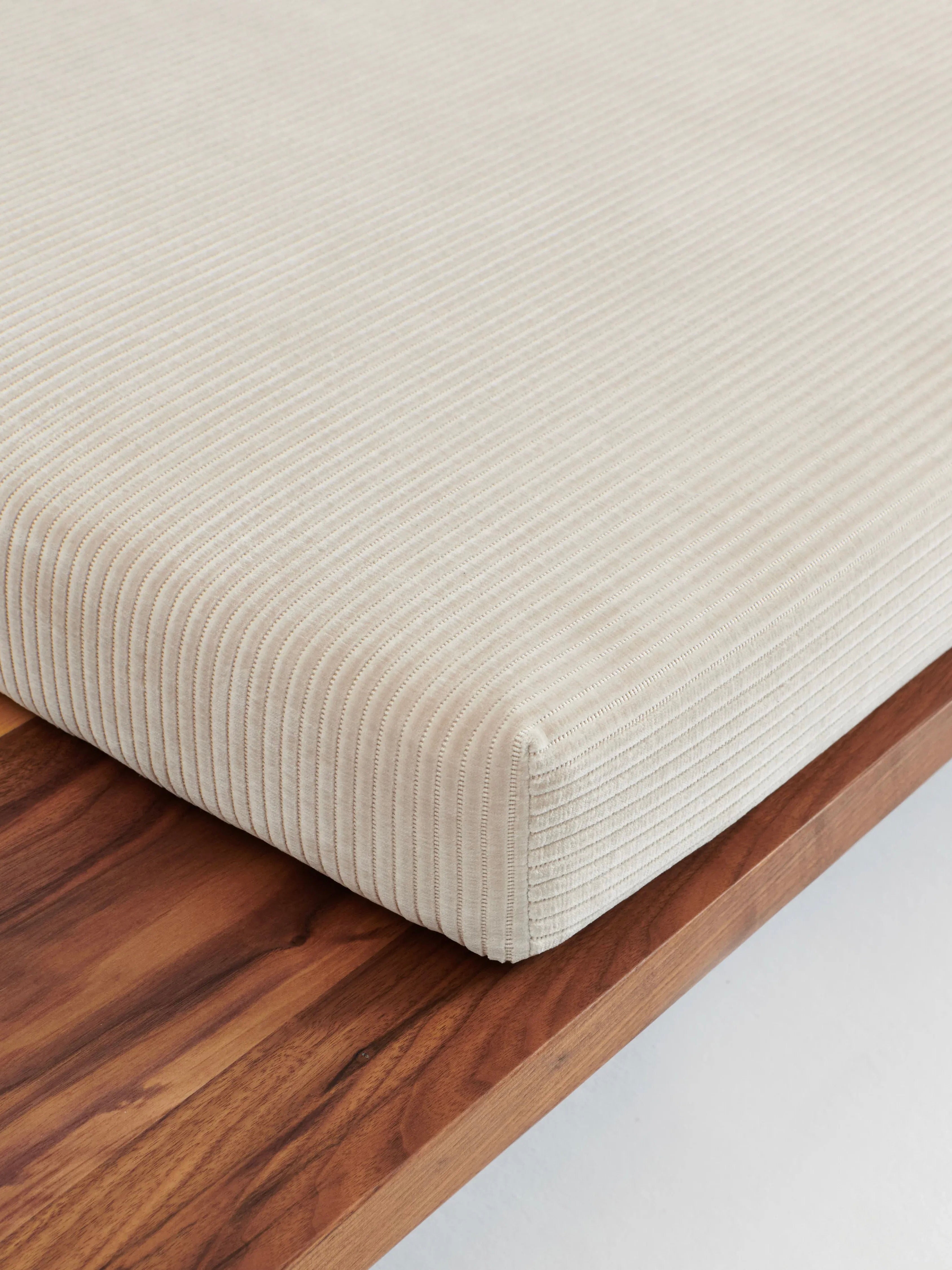 A close-up image of a neatly tucked beige, ribbed textured fabric cushion on the smooth solid walnut surface of the Nazaré Daybed L Shape by Project 213A. The wood shows natural grain and rich brown tones, while the handmade cushion from Portugal adds elegance and craftsmanship to any space.