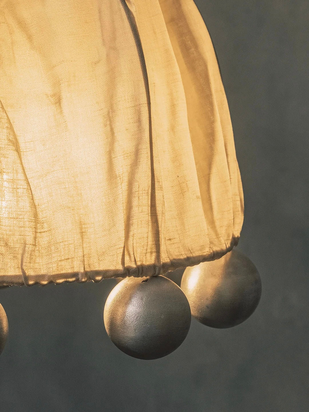 A close-up of the Kaytar "ALTER" Pendant Light lampshade reveals a warm, soft glow. The edge is adorned with three spherical decorations made from sand-cast metal, set against a textured wall background.