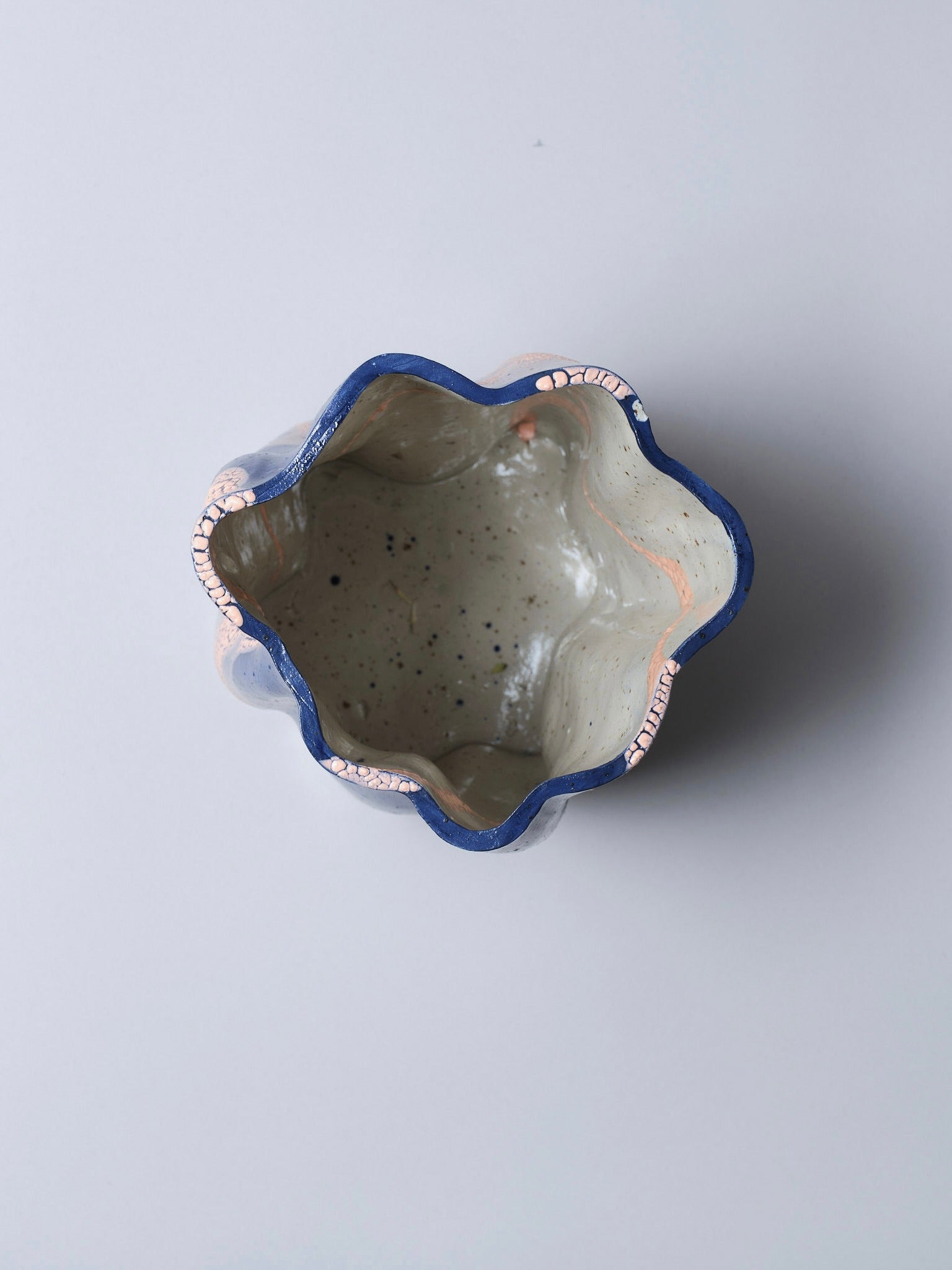 A top view of Astrid Öhman's Swirly Vase Blue and Peach shows a handmade stoneware piece with a wavy opening and speckled interior. It features a striking dark blue rim with hints of pink, elegantly set against a light grey background.