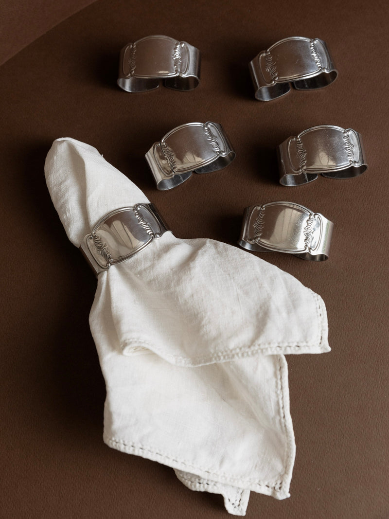 A set of 6 Stainless Steel Napkin Rings 50s by Bottega Jacobs rests on a brown surface, with one ring elegantly holding a folded white cloth napkin. The rings showcase engraved designs, adding a sophisticated touch to the table setting, ideal for an Abert Nicrosil-inspired dining experience.