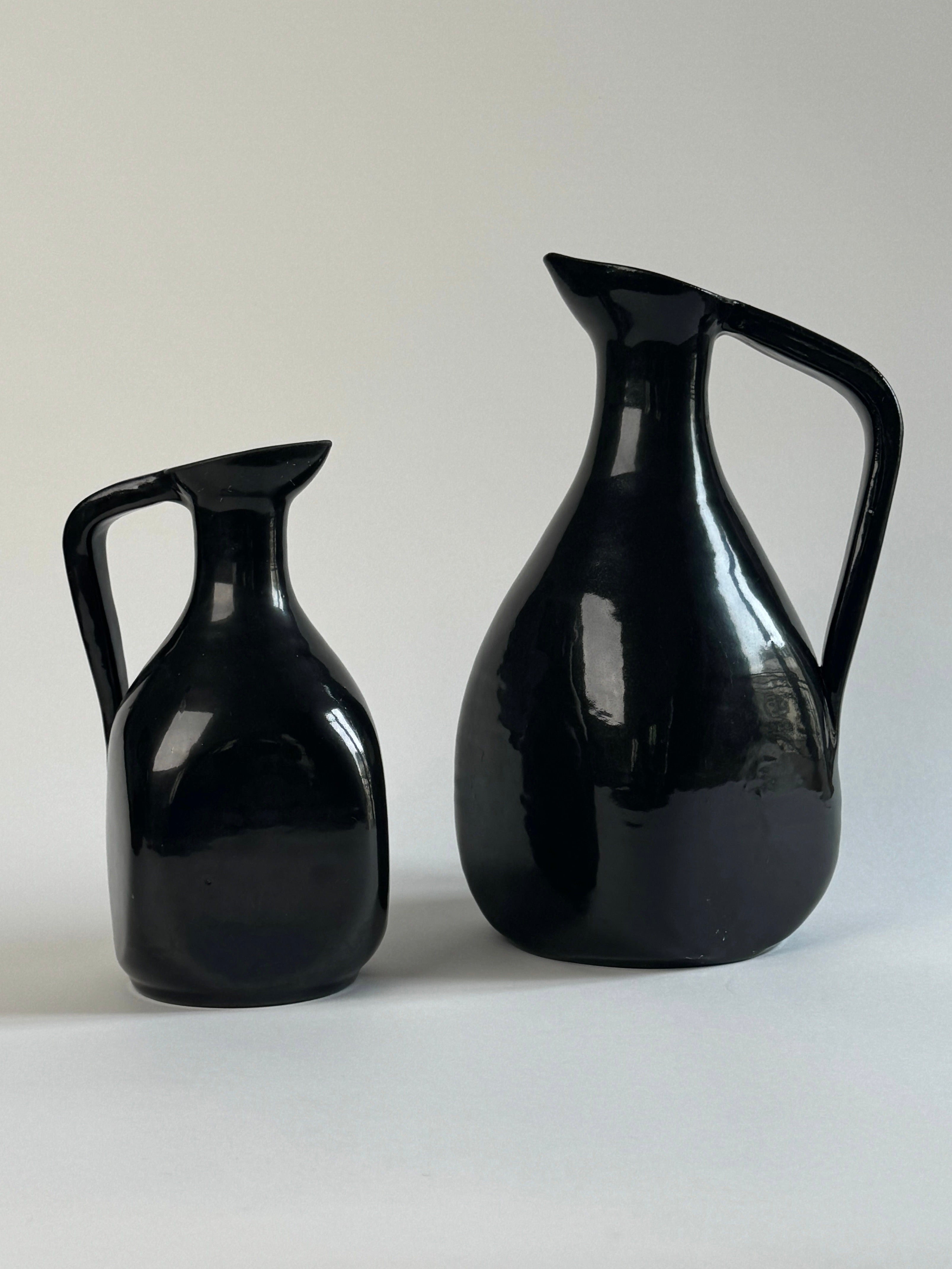 Two black glazed ceramic carafes from Figuresdesign are placed against a neutral background. The carafe on the left is smaller, with a more angular form, while the larger carafe on the right has a rounder, more bulbous shape. Both feature large handles and curved spouts, reminiscent of vintage French pottery from Accolay France.