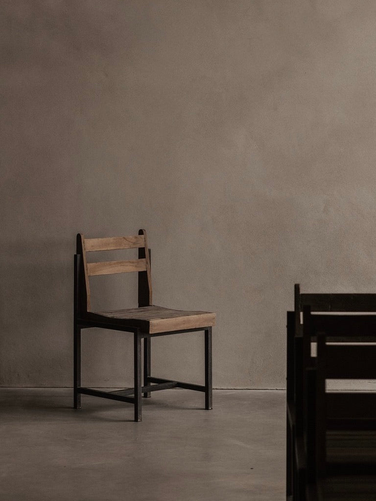 An authentic Pierre Jeanneret PJ-SI-65-A chair by Bicci de' Medici, made from teak wood slats, sits against a plain beige wall in a minimally adorned room with a concrete floor. Soft lighting casts gentle shadows, creating an atmosphere of simplicity and calmness.