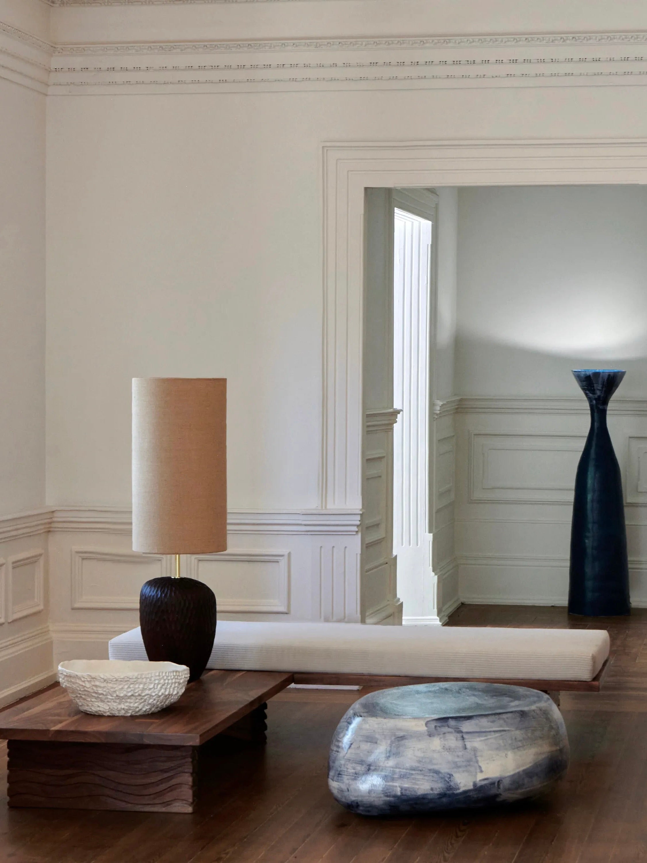 A minimalist room with white walls features a low wooden coffee table adorned with a beige lamp and a decorative bowl. The Nazaré Daybed L Shape by Project 213A, known for its modern design and comfort, sits nearby. A large, blue vase is visible in the adjacent hallway through the rectangular doorway, adding a touch of color to the serene space.