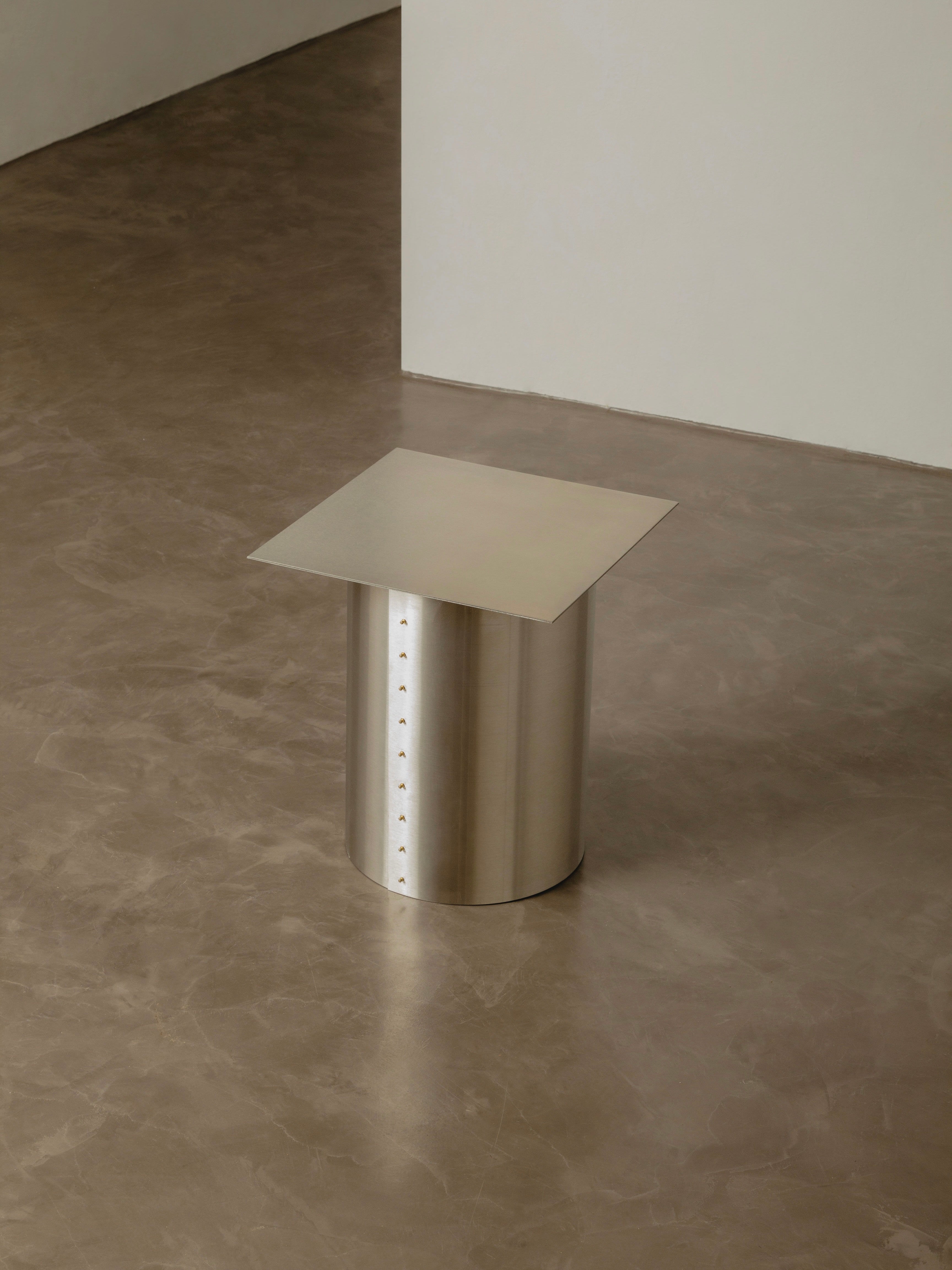 The "Plataforma" Side Table by Pedro Avila, featuring a minimalist metallic design with a cylindrical base and square top, stands on a polished concrete floor against a cream-colored wall, subtly echoing the sleek design seen in today's technological era.