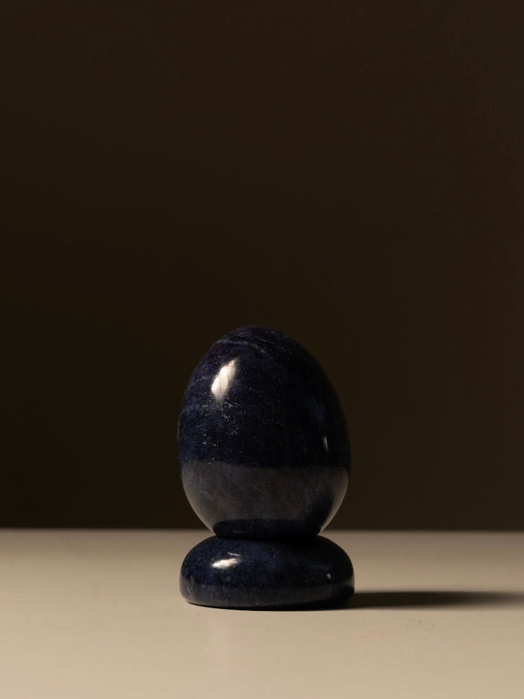 The Marble Egg Sculpture by Les Objets Amsterdam showcases a dark blue, polished Lapis marble egg standing upright on a matching round base against a plain, dark backdrop. The decorative piece is side-lit, casting a soft shadow on the light-colored surface below.