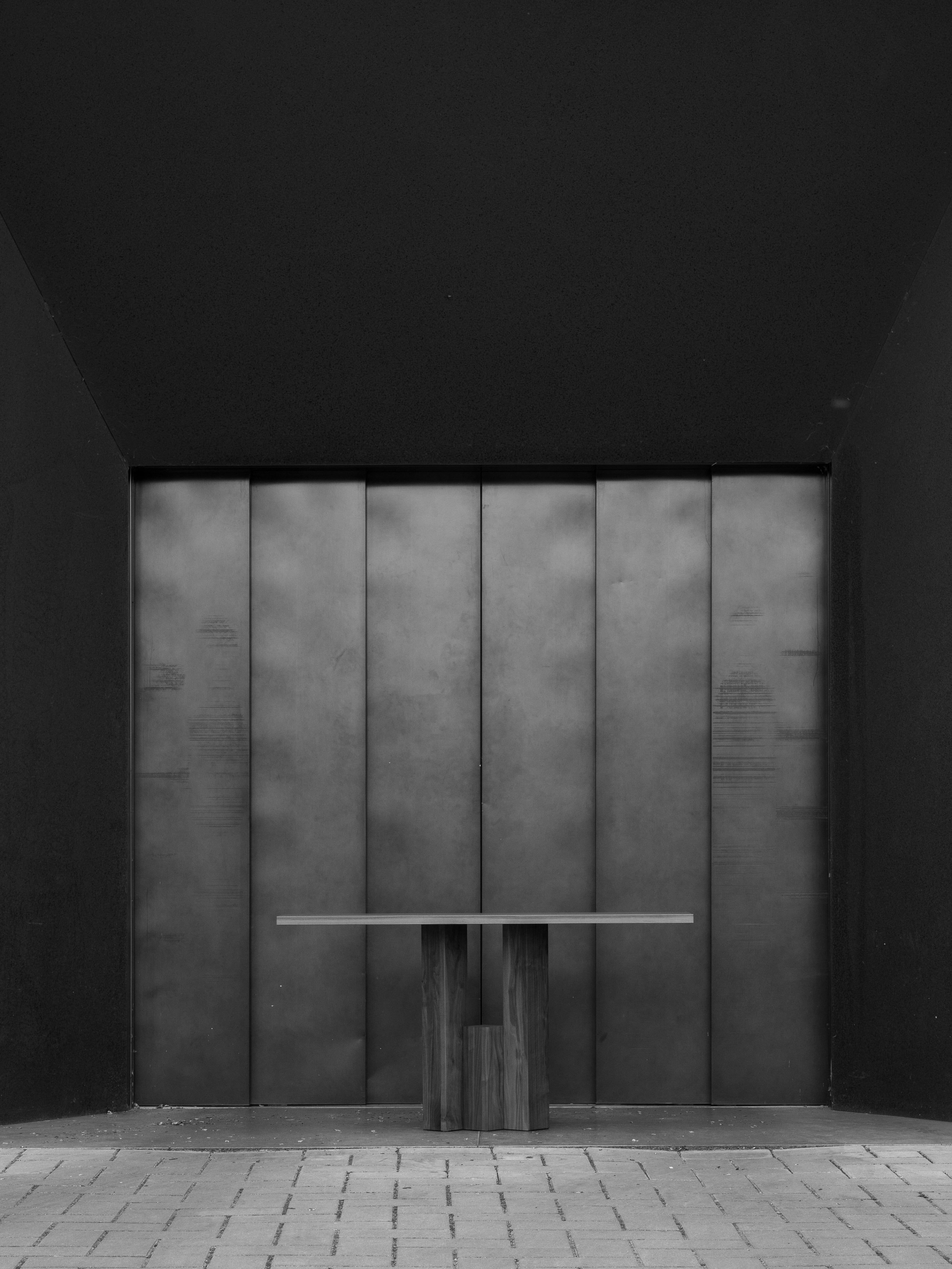 A minimalist, modern industrial-style door in a dark frame reflects Brick Expressionism. In front is a Mokko Fenestra Console featuring a wooden base and light top. Rectangular stones pave the ground.