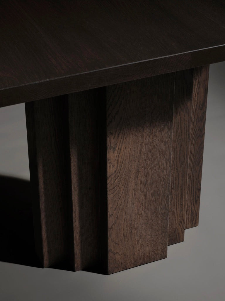 A close-up of the Brut Slim Table by Mokko showcases its minimalist and modern design, with a thick rectangular European oak top and sturdy geometric legs. The visible wood grain casts a soft shadow on the floor.