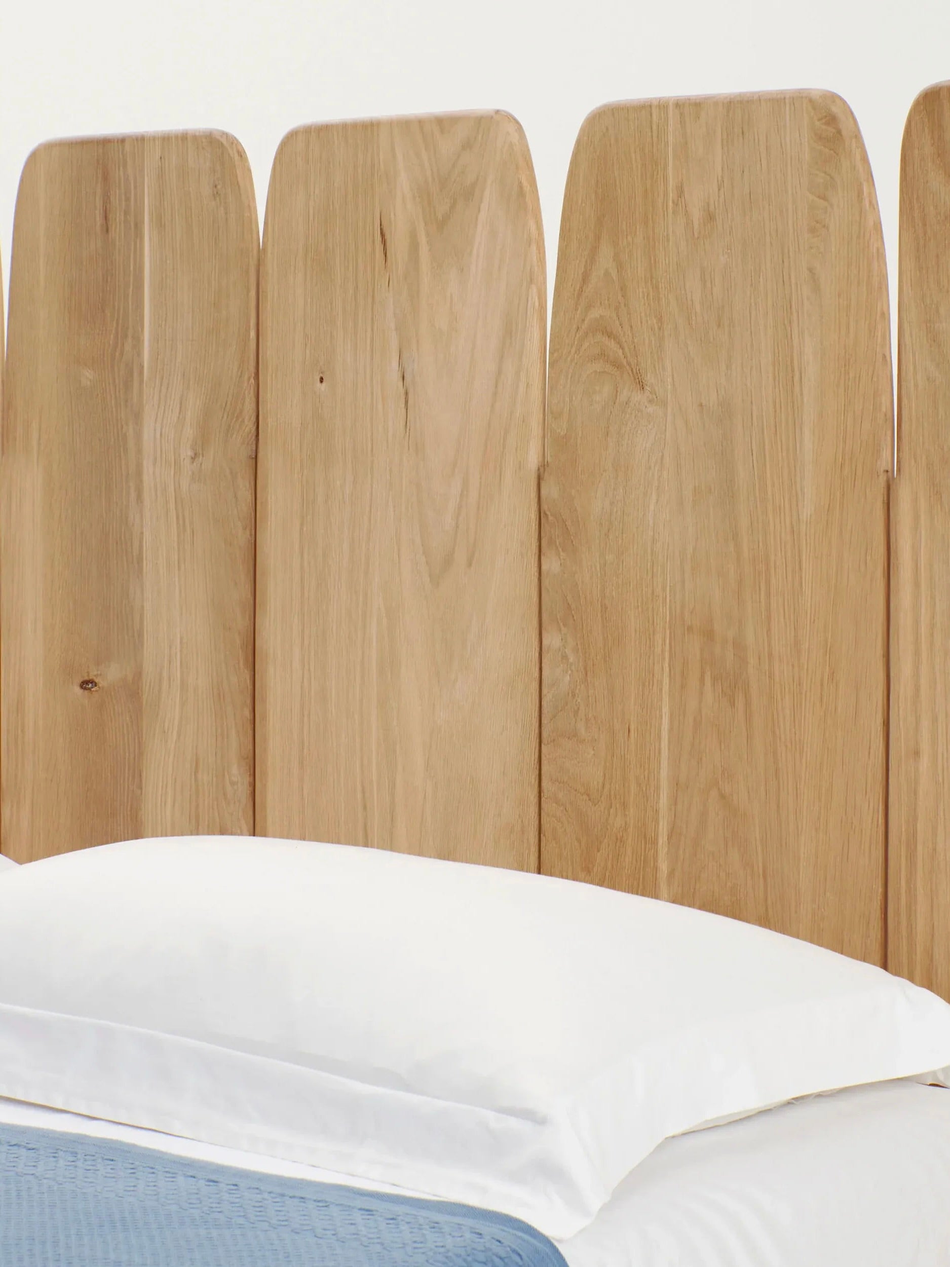 The Madeira Oak Headboard Medium by Project 213A features a natural finish and an irregular top edge that exudes a rustic charm. It is elegantly attached to a bed adorned with white pillows and sheets, complemented by a blue blanket.