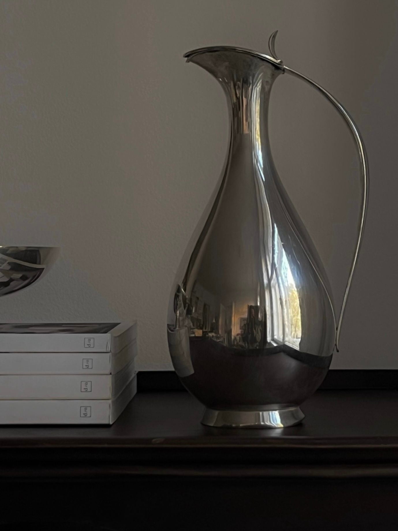 A tall, elegant Mid-Century Silver Water Pitcher by Médecine, reminiscent of a rare 1950s water jug from Finland, sits on a dark wooden surface next to a stack of four white books. The scene is softly illuminated, capturing subtle reflections on the pitcher's surface and creating a serene atmosphere.