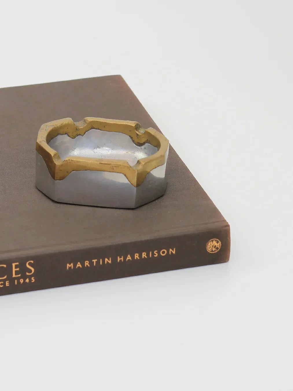 A Brutalist Ashtray by Boga Avante Shop rests on a hardcover book with the spine text "Martin Harrison." The book's cover is brown, complementing the ashtray's abstract, organic shape that embodies a touch of Brutalist design. The background is a plain white surface.