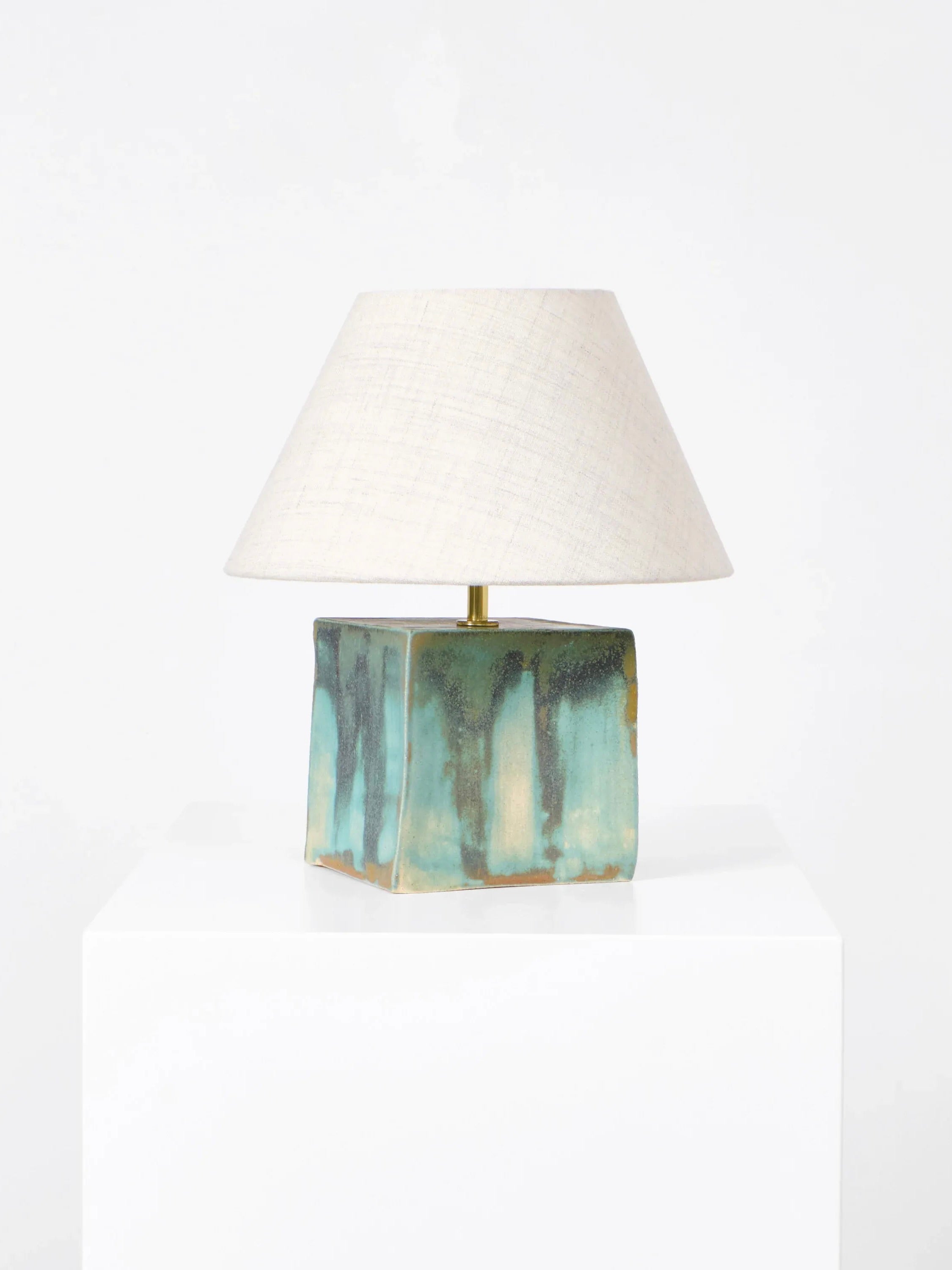 The Arouca Table Light by Project 213A features a handmade square ceramic base with a stunning blue and green glaze. It's topped with a cream-colored conical fabric shade and rests on a white pedestal against a plain background.