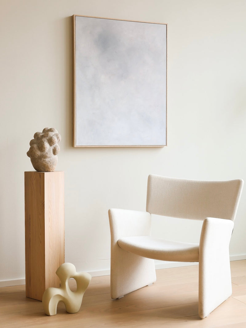 A minimalist room with a beige armchair, two abstract sculptures, and "Eos" by A Nesting Place, a large rectangular acrylic on canvas from 2024, adorning a light-colored wall. One sculpture rests on a wooden pedestal while the other sits beside the armchair. The overall color scheme is neutral, enhanced by the oak frame of "Eos.