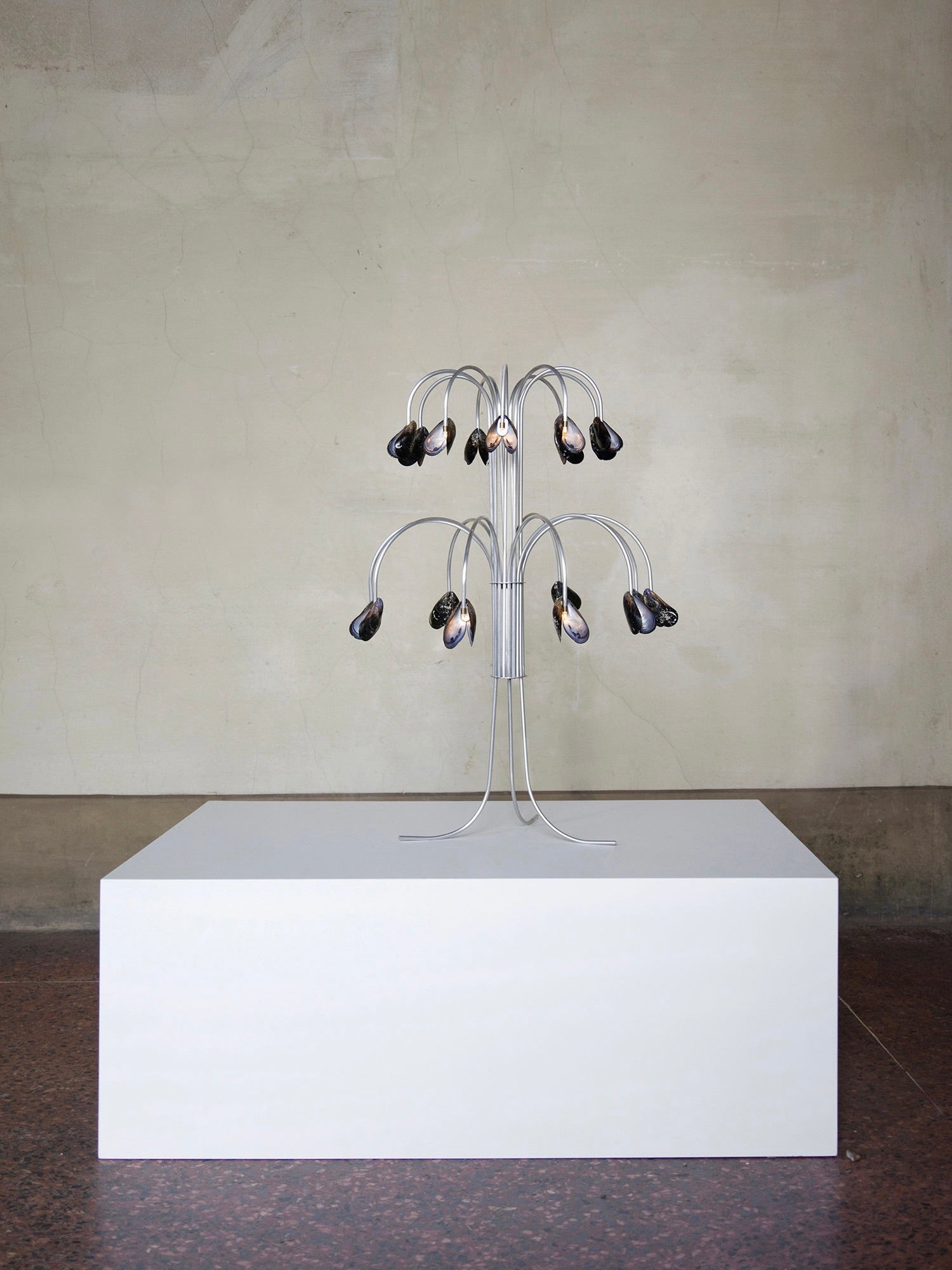 The Shell Light by Touch With Eyes is displayed on a white rectangular pedestal against a plain wall, featuring a design that resembles a metal tree sculpture with multiple curved branches, each ending in black and metallic elements that exude authenticity.