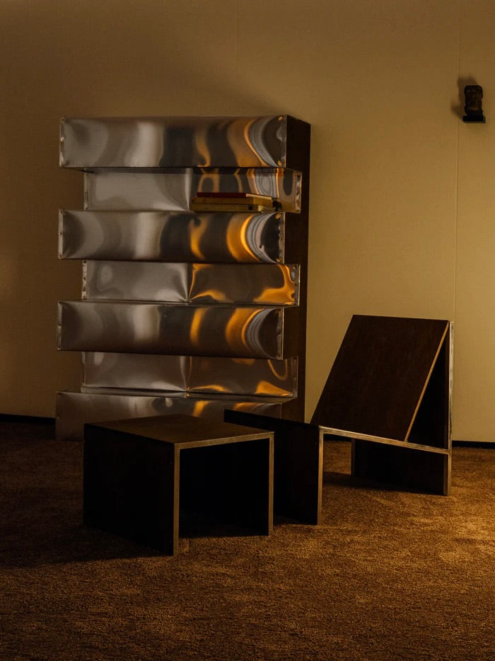 A minimalist room features a brown chair, two matching side tables, and The Aluminum Bookshelf by Studio HAOS against a beige wall. Warm light casts subtle reflections across the shelving unit’s metallic surface.