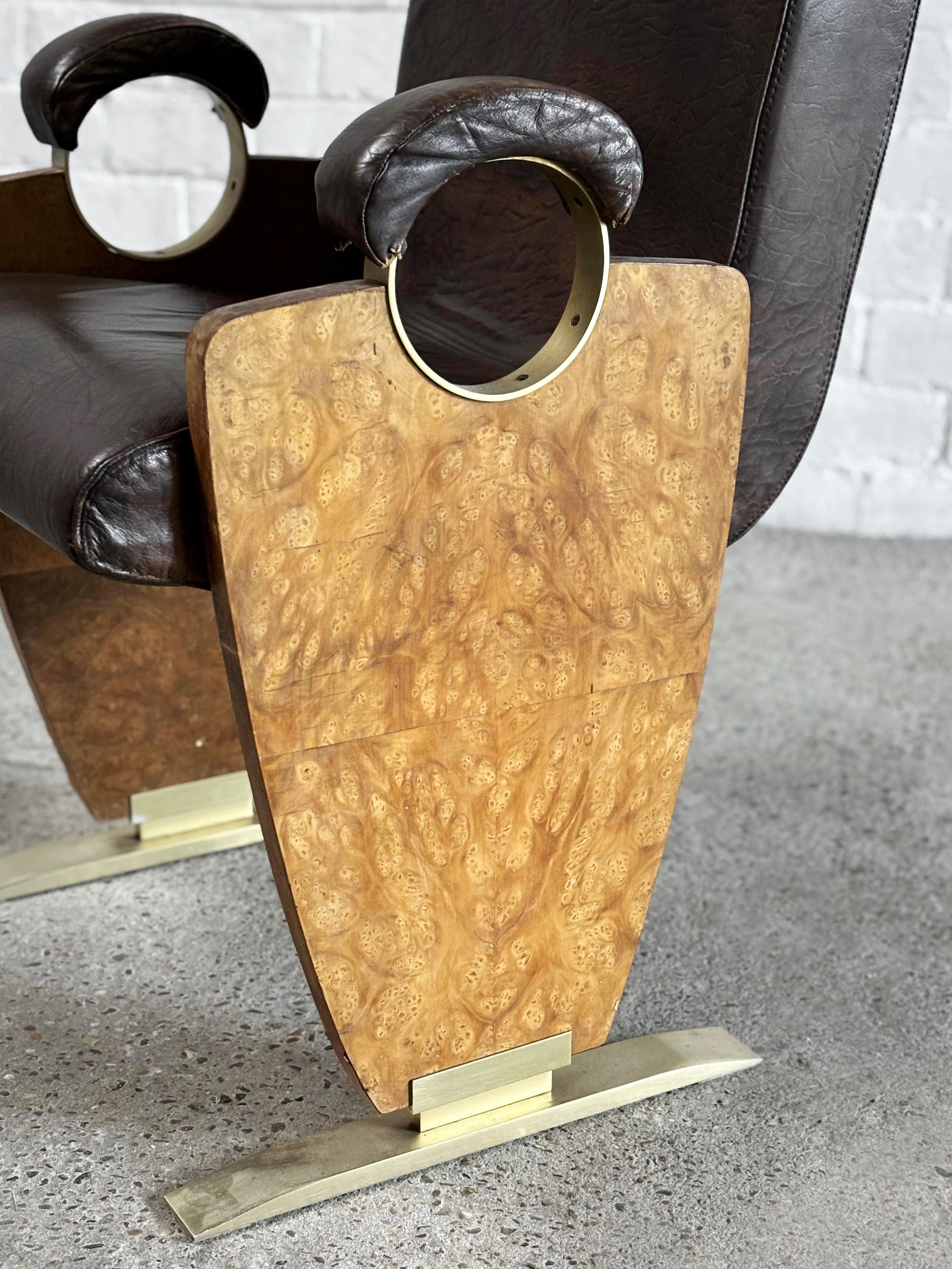 Close-up of an Art-Deco Style Side Chair by Introverso from the 1970s, showcasing dark skai leather upholstery, a unique burl walnut wood panel with ornate patterns, metallic circular accents, and a chrome base. It stands on a light concrete floor against a white brick wall.