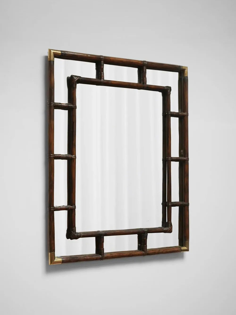 The Brown Rattan and Brass Mirror from Barracuda Interiors, inspired by the 1970s, features a wooden frame crafted to resemble bamboo with textured rattan details. The interior frame is suspended within the outer frame for a layered appearance. Mounted on a plain white wall, it elegantly combines natural warmth with minimalist simplicity.