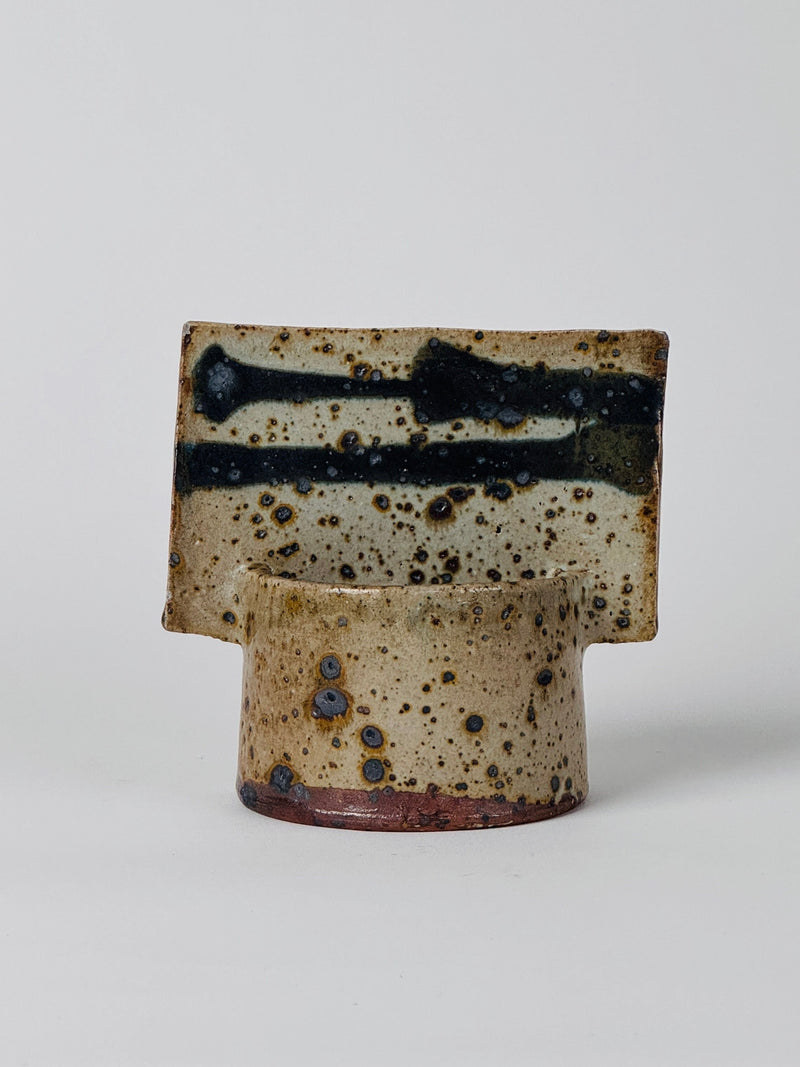 The Geometric Vase by Gustave Tiffoche from Figuresdesign features a cylindrical ceramic base paired with a distinctive, flat, rectangular top that evokes the essence of pyrite stoneware. Its speckled brown and beige surface with dark blue streaks and spots imparts a rustic, artisanal appearance. The vase stands against a plain white background.