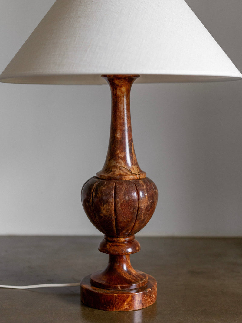 A Spigel Large Italian Alabaster Stone Lamp, adorned with a white conical shade and featuring a brown, marbled ceramic base in an elegant neoclassical sculpted design, sits on a gray surface. The lamp is connected with a white cord trailing to the side.
