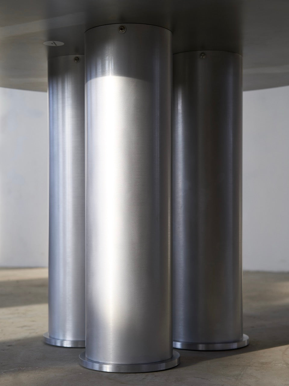 Three large cylindrical metal pillars on a concrete floor support a flat round surface above, creating an industrial setting akin to the Elephant Tripod Table by Frederik Fialin, with soft lighting casting shadows.