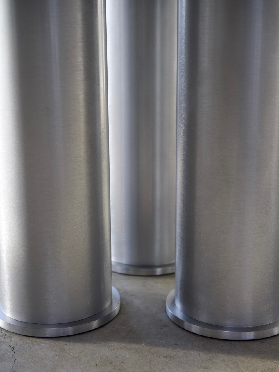 Close-up of tall, cylindrical metal columns with a smooth, brushed finish on a concrete floor in Frederik Fialin's studio. The lighting accentuates the texture and reflects beautifully, ideal for complementing an aluminium dining table or an Elephant Tripod Table by Frederik Fialin.