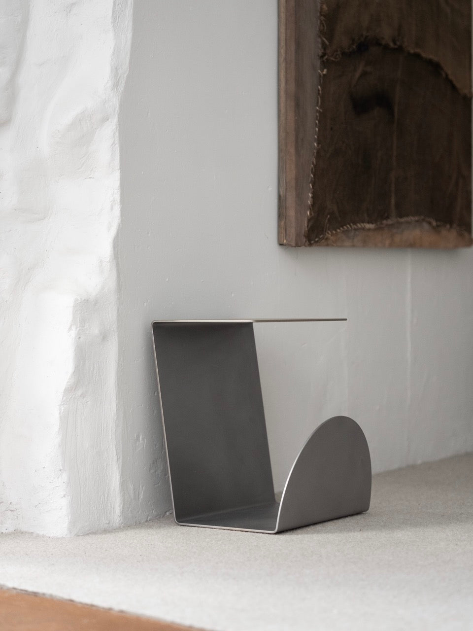 A modern minimalist stainless steel side table by KØGE Design, featuring a sleek gray finish, sits against a textured white wall beneath rustic artwork, perfectly complementing an elegant contemporary interior with its geometric design.