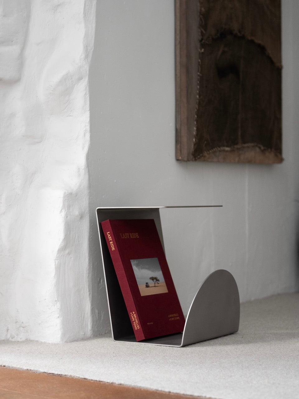 A minimalist KØGE Design stainless steel side table holds a red book with a tree on the cover, positioned on the floor against a textured white wall, beside hanging artwork. The contemporary interior exudes a clean, modern aesthetic.