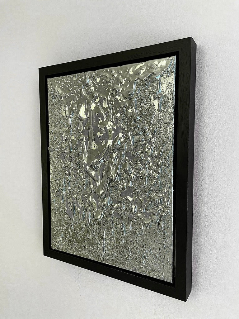 The "Almost a Mirror" Sculpture by John Nordenstein is showcased in a black rectangular frame on a plain white wall. This unique piece features a textured silver abstract design with a reflective, metallic surface and intricate patterns that resemble liquid metal or melted wax, evoking organic shapes.