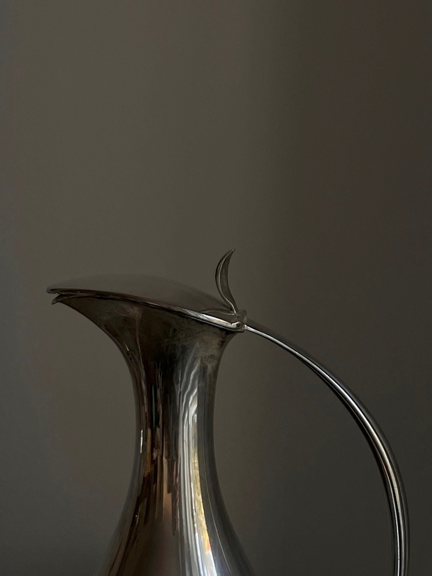 Positioned against a dark, neutral background is a rare Mid-Century Silver Water Pitcher by Médecine, originating from 1950s Finland. The pitcher features a narrow spout and curved handle, and its sleek, reflective metallic surface elegantly catches the light, accentuating its smooth design.