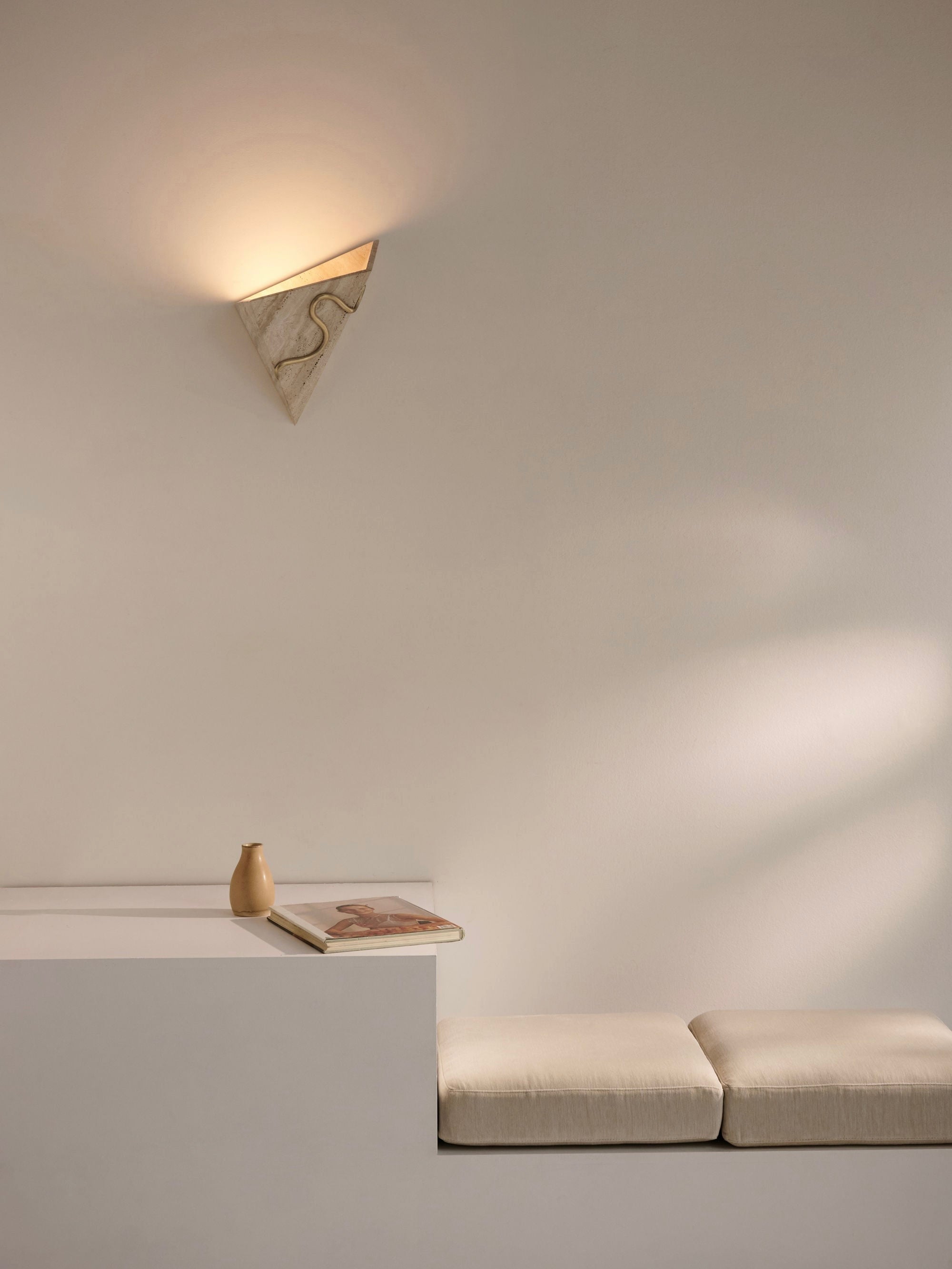 A minimalist interior featuring a white wall with a customizable finish and the Mariza Galani Therapeutic Wall Light 1, emitting a soft glow. Below it is a low, white bench with two white cushions. A small beige vase and a book are placed on a sleek, white table adjacent to the bench.