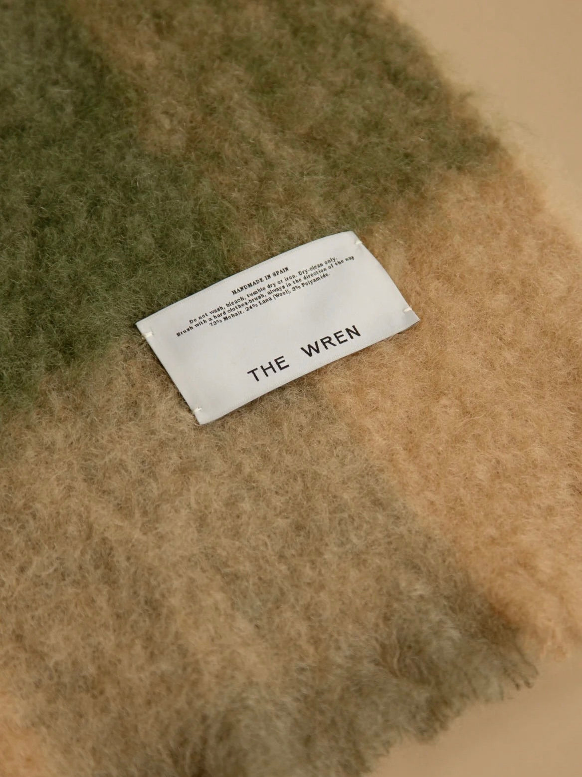 The "Arbor no. 2" Throw from The Wren is a soft, luxurious mohair blanket featuring shades of green and brown. A distinctive white label prominently displays "THE WREN," and the eco-friendly fabric provides both comfort and elegance.