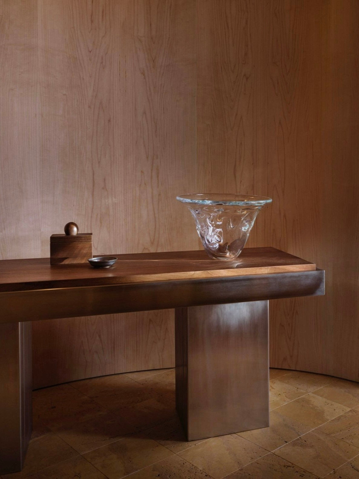 A minimalist interior features the Studio Vraco Console Table 02 adorned with a decorative glass bowl, a small wooden box, and a metal dish, all elegantly set against curved wooden paneling. The design captures the contemporary aesthetic while subtly incorporating Bauhaus furniture principles.