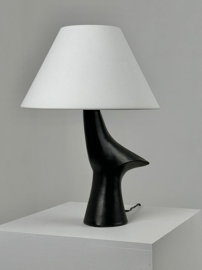 The Zoomorphic Ceramic Lamp by Figuresdesign features a sleek black base that resembles an abstract sculpture, topped with a minimalist white conical lampshade. Made from black glazed ceramic, it is displayed on a white pedestal against a plain gray background, capturing the sophisticated design ethos of 1960s France.