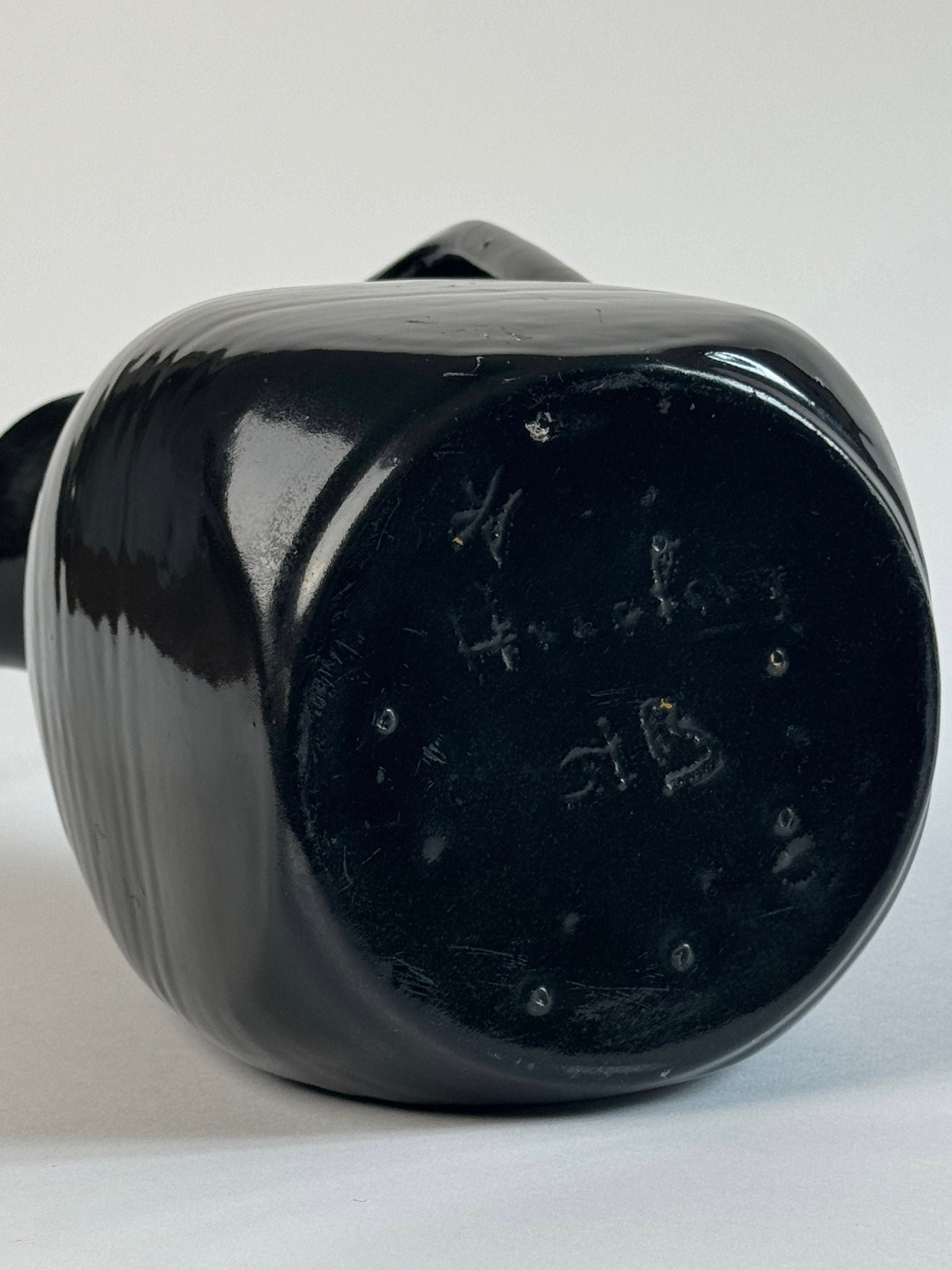 A close-up shot of the base of a Pair of Accolay Ceramic Carafes, glazed in black and featuring some indiscernible markings etched into the bottom. The carafes by Figuresdesign are positioned horizontally on a plain, light-colored surface, reminiscent of vintage French pottery.