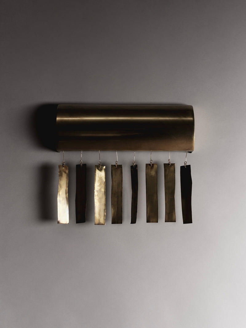 The Ombia Studio Quimbaya Sconce is a handmade brass wall fixture featuring eight dangling metal rectangles in gold and bronze shades. Mounted on a smooth gray wall, it casts gentle shadows with its cylindrical base design.