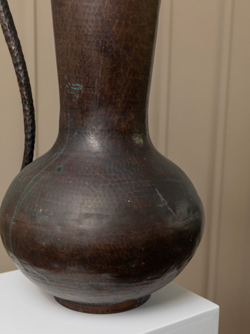 A close-up of the Giant Vintage Metal Jug - Italy 1950s by Bottega Jacobs showcases a large, handcrafted dark bronze vase with a textured surface reminiscent of a vintage metal jug. Featuring a bulbous base and narrow neck, it exudes timeless charm on its white pedestal against a neutral background, echoing the artistic flair of Italy in the 1950s.