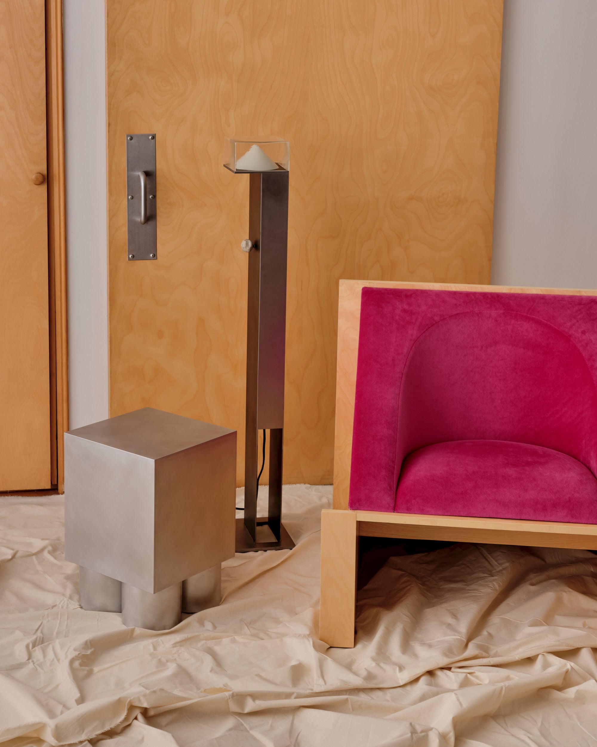 A modern interior showcases a Known Work 'Perceptions' Armchair in pink velvet, paired with a metallic cube-shaped stool and a tall floor lamp. The minimalist design is completed by a wooden wall and door backdrop, with cream fabric draped elegantly on the floor.