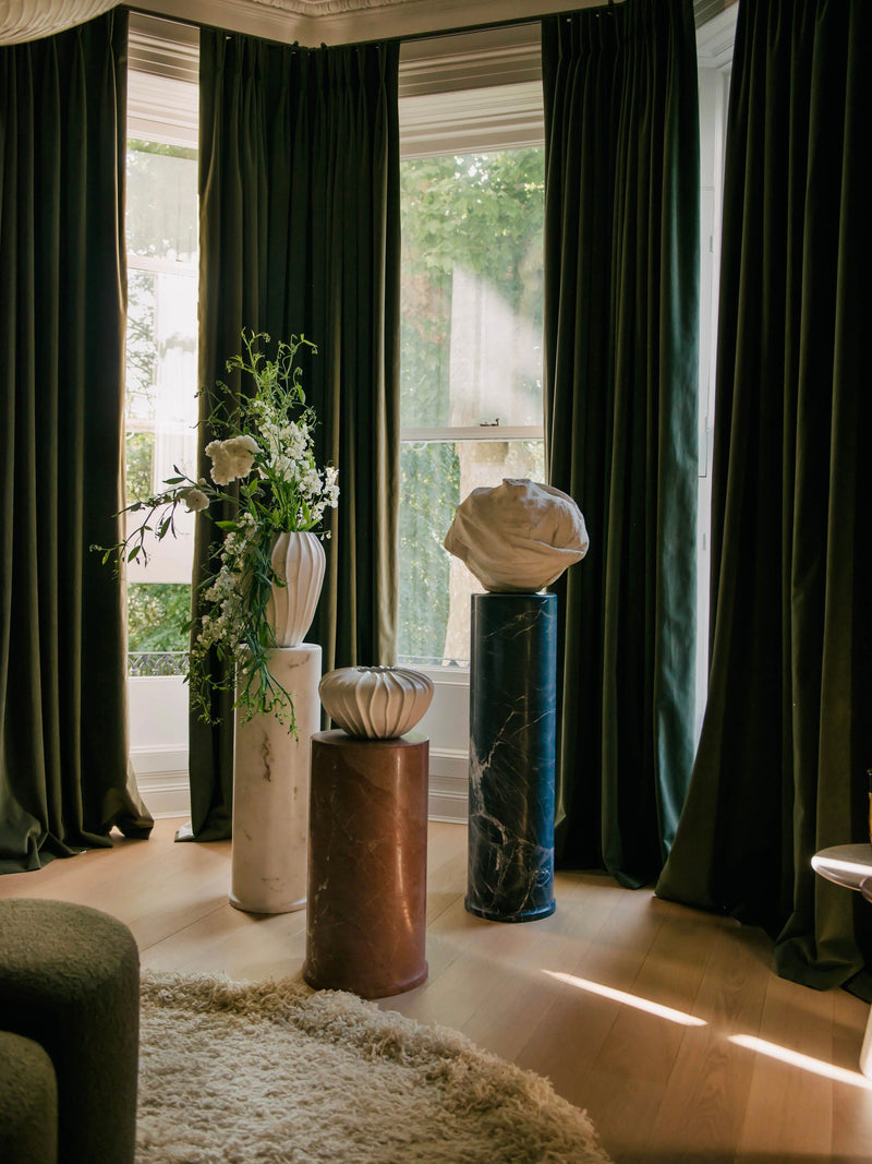 A cozy, elegantly decorated room features four tall Wide Cloak Vases from Ransom & Dunn, standing proudly on a wooden floor. Dark curtains are partially drawn, allowing sunlight to stream through a large window and cast soft shadows on the vases and floor. Each piece is handcrafted in Portugal.