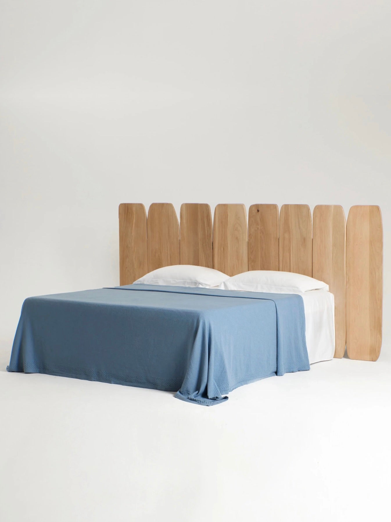 Presenting the Madeira Oak Headboard Medium by Project 213A - a minimalist bed featuring a distinctive headboard crafted from vertical, boat-shaped slats. The meticulously made oak wood frame is complemented by a neatly arranged blue blanket and two white pillows. Set against a plain white backdrop, this customizable bed frame highlights its clean lines and natural wood tones elegantly.