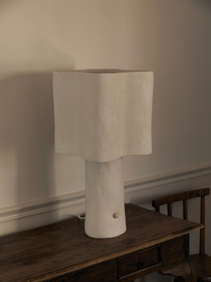 The Freeform Table Lamp by Studio Tristan Kallas sits on a wooden table against a light wall. Its minimalist white design and unique wavy shape softly illuminate the room, with a wooden chair partially visible beside the table.