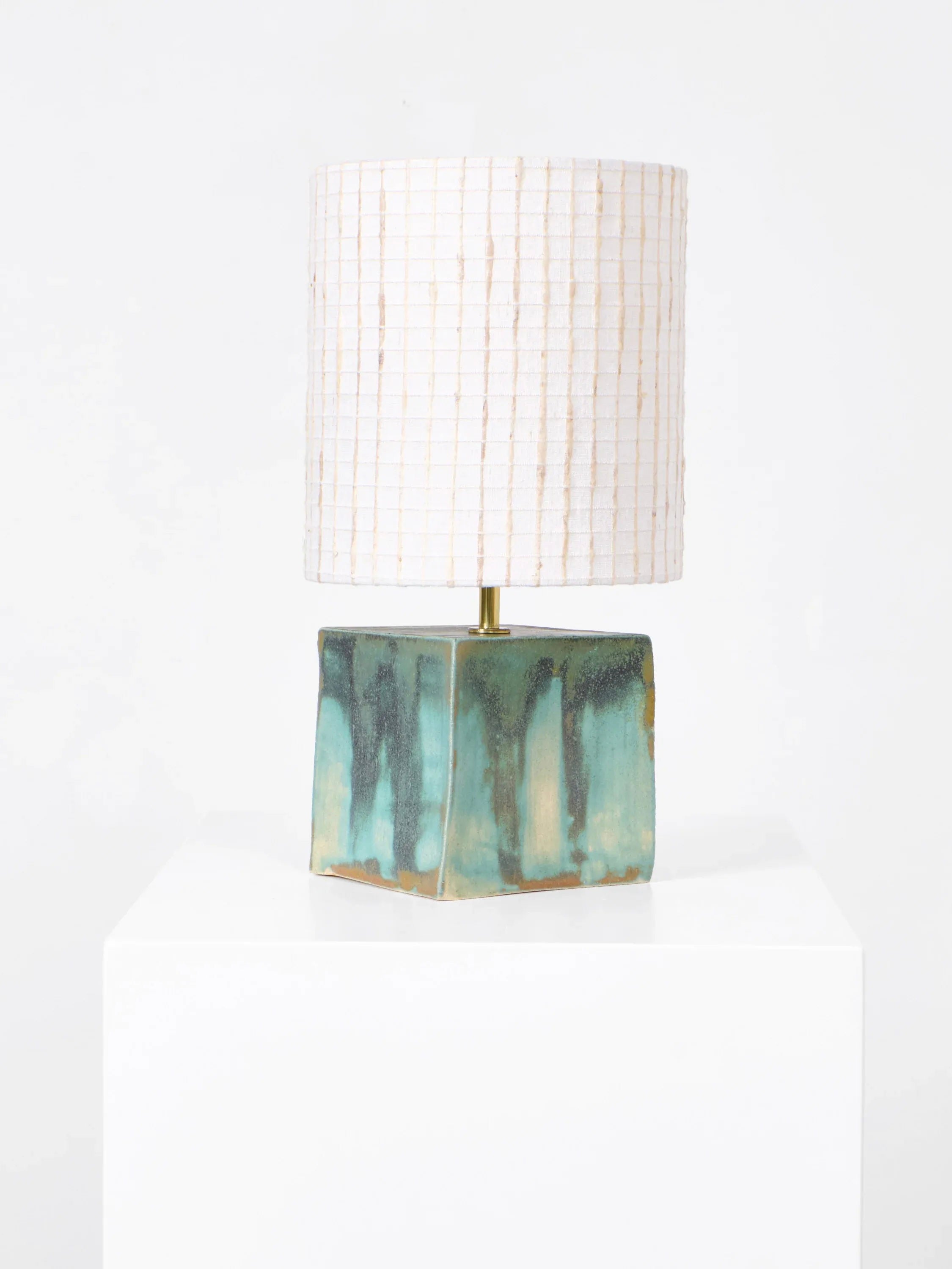 The Arouca Table Light by Project 213A features a handcrafted ceramic base in green and brown, topped with a cylindrical white fabric shade with a subtle grid pattern. This artisanal piece elegantly sits on a white pedestal against a plain backdrop.