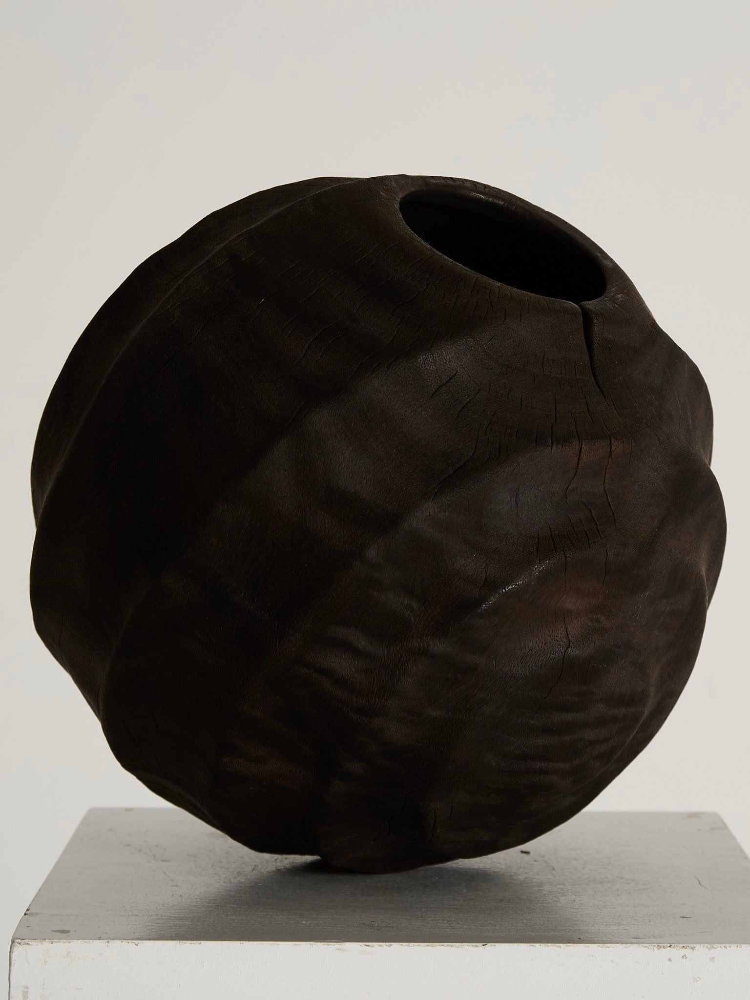 The Ancien et Jolie Hardwood Sculptured Vessel, inspired by British 1970s design, features a dark, textured ceramic with a spherical shape and small opening. Its subtly swirling patterns and uneven finish make it ideal for displaying dry stems with minimalist elegance on a white platform.