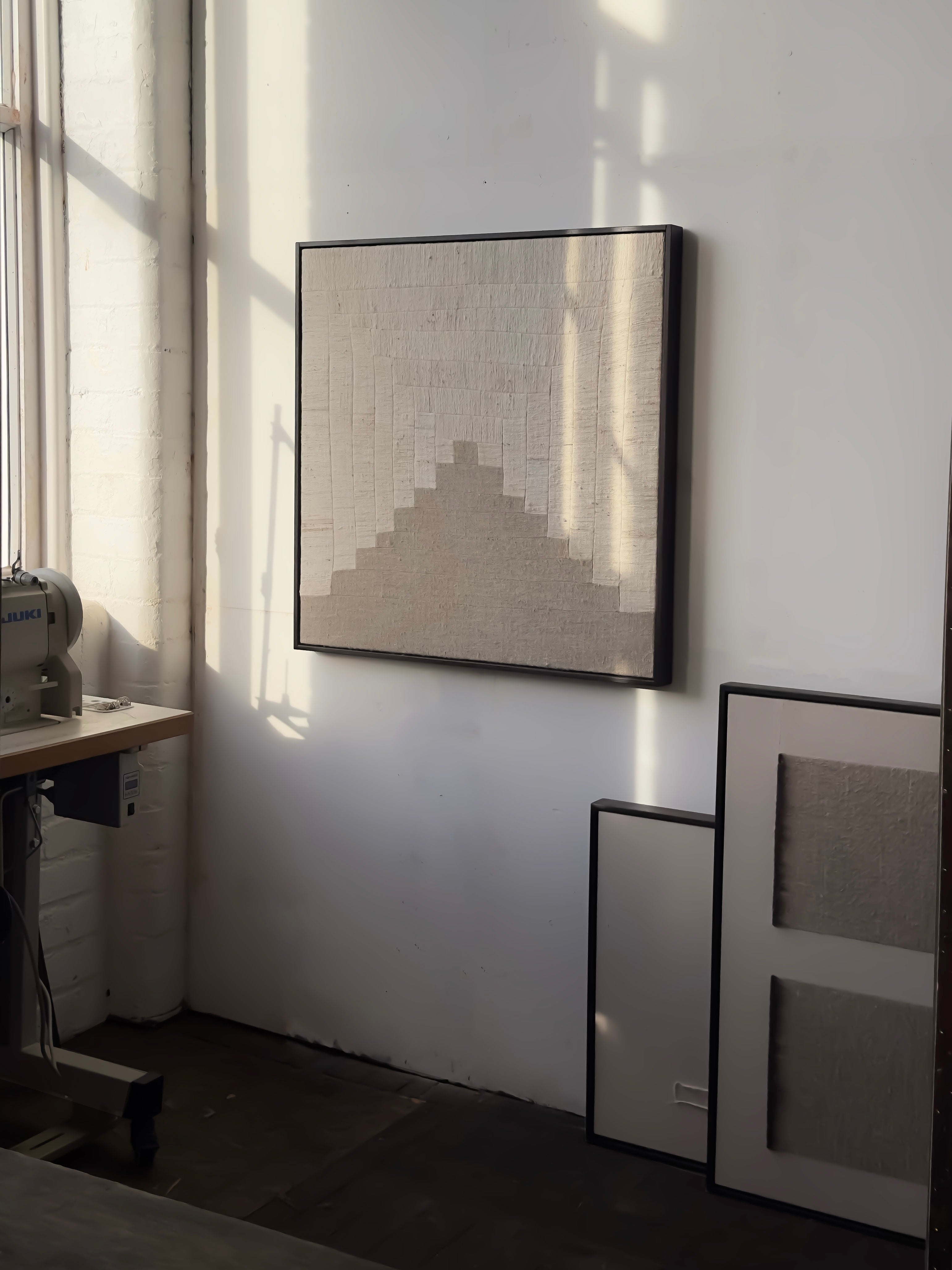 A room corner features minimalist art: "People's Hill" by Bec Kirby, a square canvas with textured geometry hangs illuminated by sunlight. Below, framed artworks lean against the wall beside a sleek solid oak desk adorned with a sewing machine.