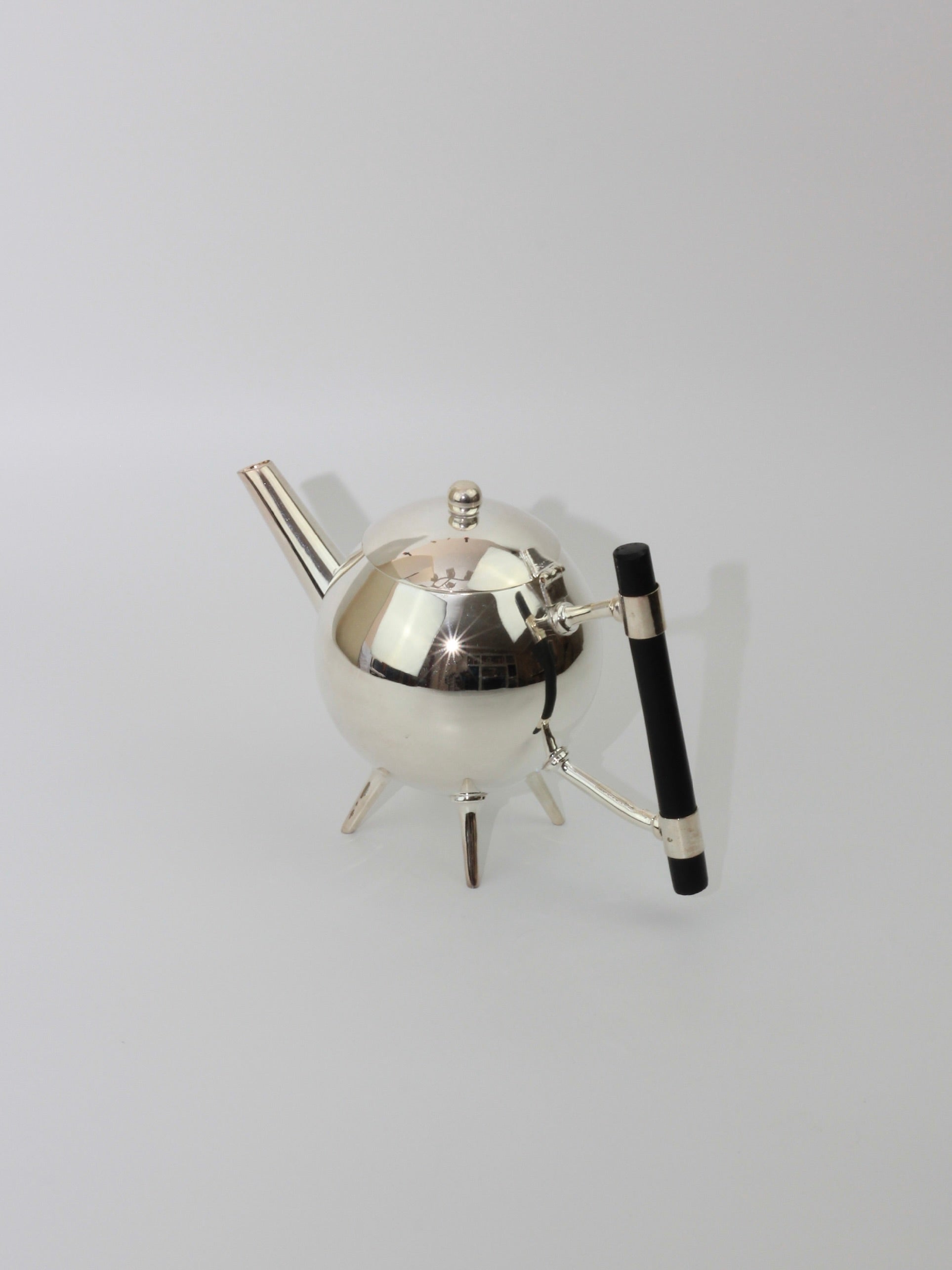 Christopher Dresser's Tea Pot, offered by Boga Avante Shop, is a sleek and modern silver-plated teapot designed with a polished spherical body that resembles a globe and stands on three small legs. It features a thin spout and an ebonized wood handle extending to the right, all set against a plain, light background—a design masterfully influenced by Dr. Christopher Dresser.