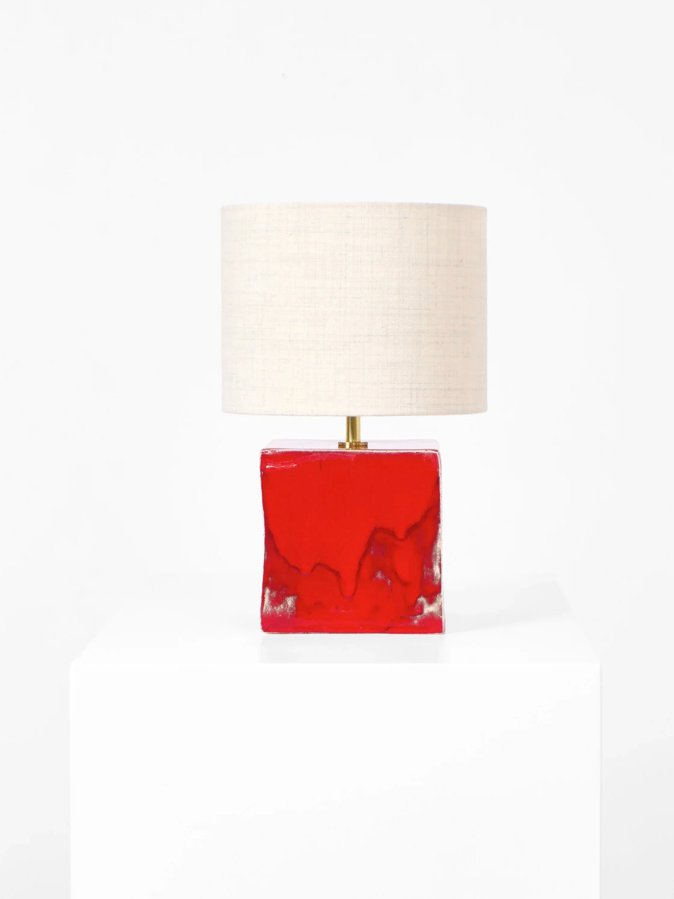 The Arouca Table Light by Project 213A, boasts a minimalist design with a rectangular red ceramic base and round white fabric lampshade, elegantly displayed on a white surface against a plain white background.