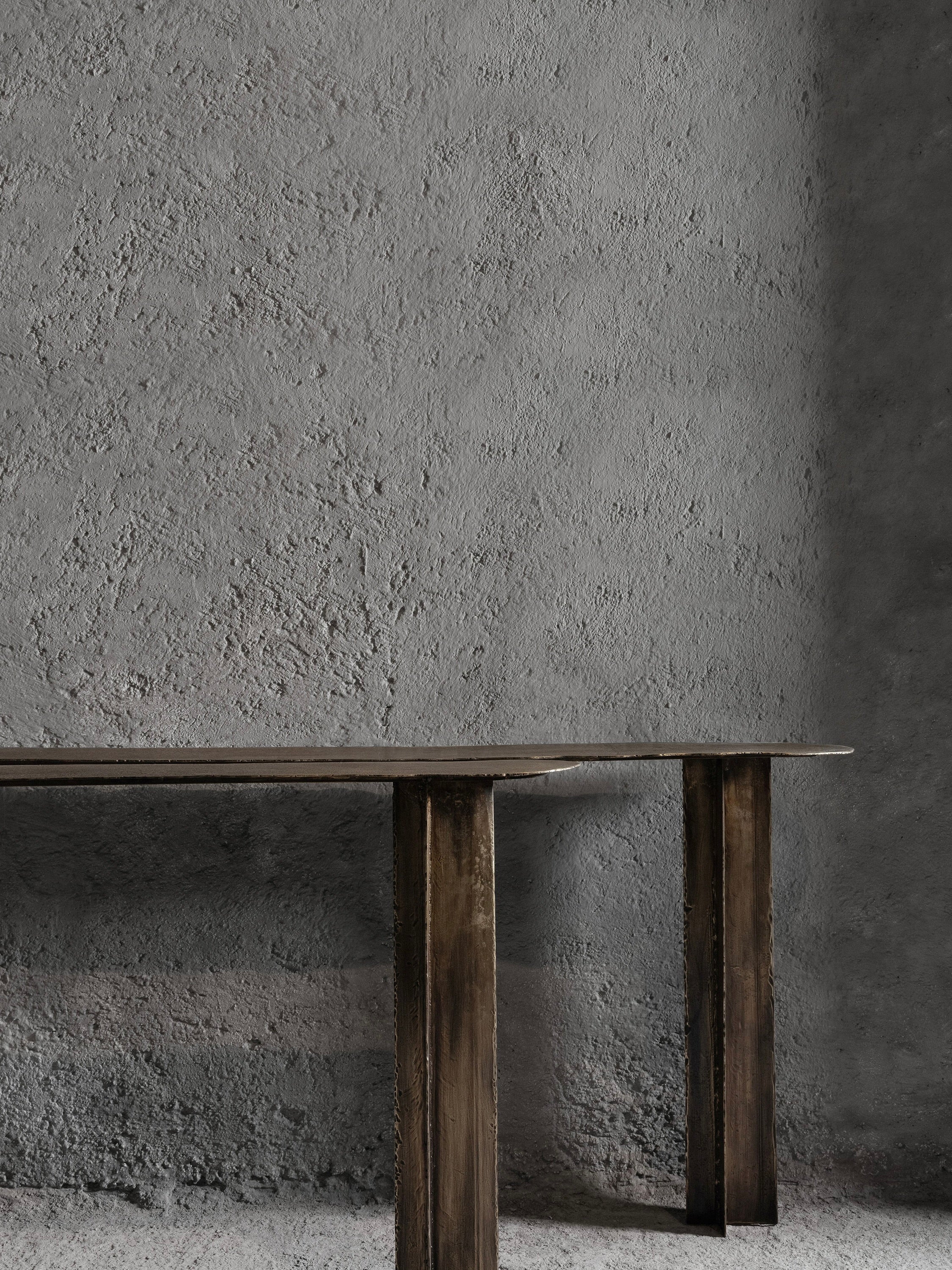 A minimalist interior features the Muisca Console Table by Ombia Studio, with its dark wood and brass-textured finish contrasting beautifully against a textured gray wall. The lighting casts soft shadows, highlighting both the wall's roughness and the table's understated elegance.